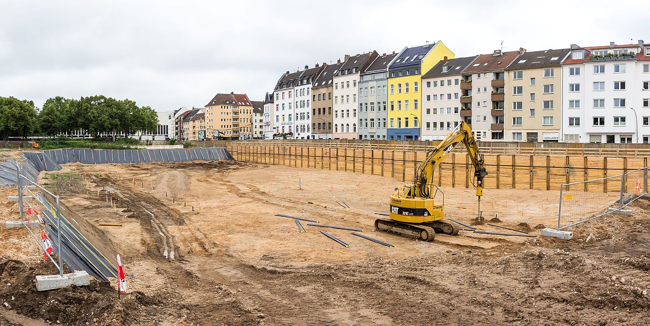 Blick auf eine sehr große Baugrube, unten ein vergleichsweise kleiner Bagger in Aktion. Hinten eine Reihe von Straßenhäusern