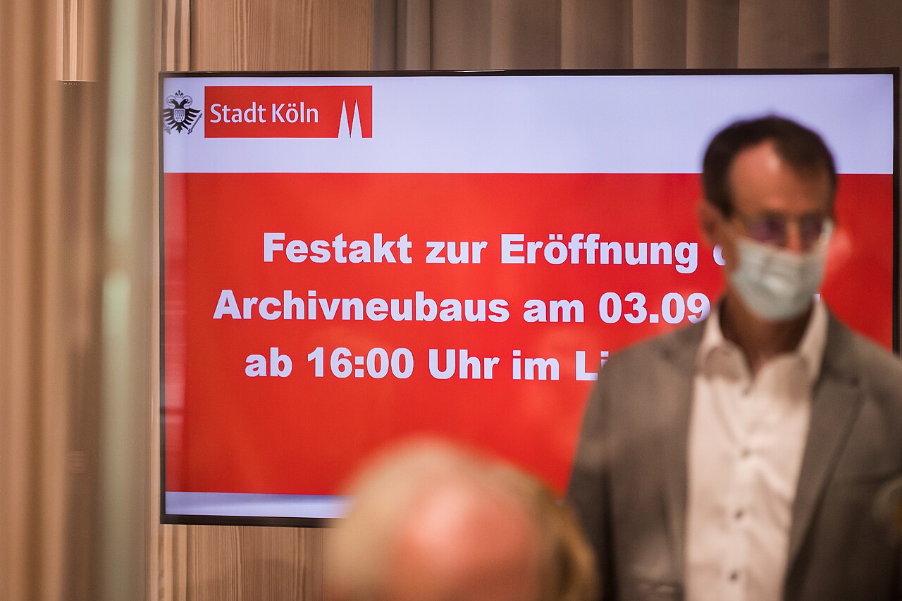 Ein großer Screen mit weißer Schrift auf rotem Grund und Stadt Köln Logo. Festakt zur Eröffnung des Archivneubaus am … davor eine unscharfe Person mit Mundschutz