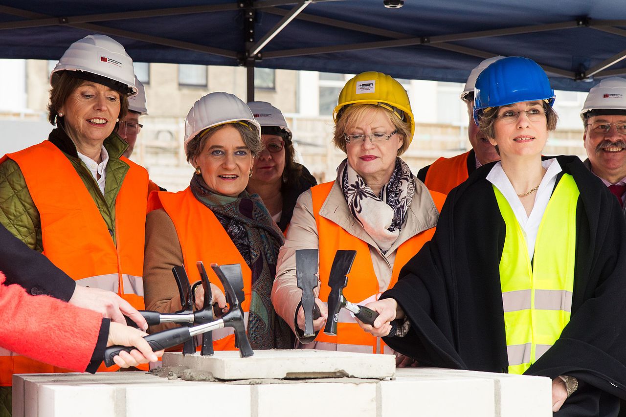 Vier Frauen mit Helmen und Sicherheitswesten auf der Baustelle, Hammer in der Hand, bei einer Grundsteinlegung