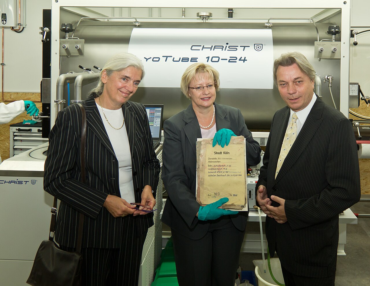 Gruppenfoto vor einer Gefriertrockungsanlage, zwei Frauen ein Mann in formeller Kleidung, die Dame in der Mitte hält ein historisches Dokument in der Hand, die mit türkisen Gummihandschuhen geschützt sind 